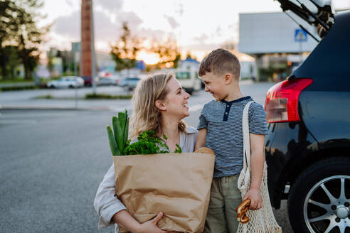 Eine junge Mutter mit ihrem kleinen Sohn hält nach dem Einkaufen abfallfreie Einkaufstüten mit Lebensmitteln in der Nähe des Autos. - HPIF04485