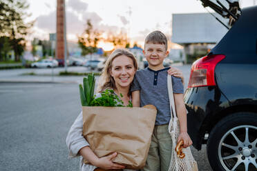 Eine junge Mutter mit ihrem kleinen Sohn hält nach dem Einkaufen abfallfreie Einkaufstüten mit Lebensmitteln in der Nähe des Autos. - HPIF04486