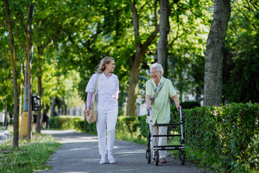 Eine Pflegerin mit einer älteren Frau auf einem Spaziergang mit Rollator im Park mit Einkaufstasche. - HPIF04499