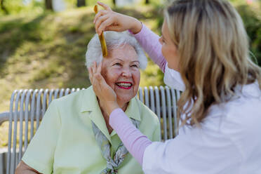 Eine Pflegekraft hilft einer älteren Frau beim Kämmen der Haare und beim Herstellen einer Frisur, während sie im Sommer auf einer Bank im Park sitzt. - HPIF04516
