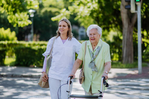 Eine Pflegerin mit einer älteren Frau auf einem Spaziergang mit Rollator im Park mit Einkaufstasche. - HPIF04517
