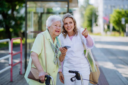Eine Pflegekraft begleitet eine ältere Frau bei einem Spaziergang im Park mit einer Einkaufstasche und hilft ihr bei der Benutzung eines Smartphones. - HPIF04519