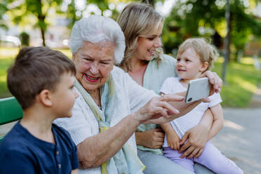 Eine Urgroßmutter, die ein Selfie mit ihrer Enkelin und ihren Kindern macht, während sie im Sommer im Park sitzt. - HPIF04527