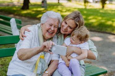 Eine Urgroßmutter, die ein Selfie mit ihrer Enkelin und ihrem Kind macht, während sie im Sommer im Park sitzt. - HPIF04528