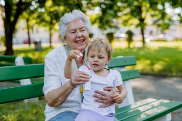 A great grandmother sitting on bench with her granddaughter and blowing soap bubbles together, generation family concept. - HPIF04530