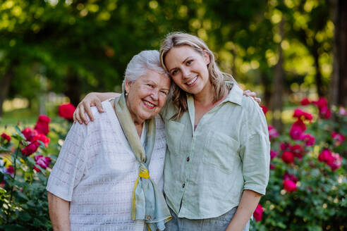 A portrait of adult granddaughter with senior grandmother on walk in park, with roses at background - HPIF04533