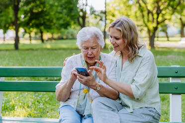 Eine erwachsene Enkelin hilft ihrer Großmutter bei der Benutzung des Mobiltelefons, während sie im Sommer auf einer Bank im Park sitzt. - HPIF04534