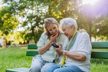 Eine erwachsene Enkelin hilft ihrer Großmutter bei der Benutzung des Mobiltelefons, während sie im Sommer auf einer Bank im Park sitzt. - HPIF04535