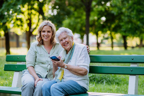 Eine erwachsene Enkelin hilft ihrer Großmutter bei der Benutzung des Mobiltelefons, während sie im Sommer auf einer Bank im Park sitzt. - HPIF04536