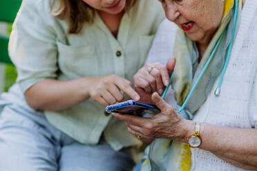 Eine erwachsene Enkelin hilft ihrer Großmutter bei der Benutzung des Mobiltelefons, während sie im Sommer auf einer Bank im Park sitzt. - HPIF04537