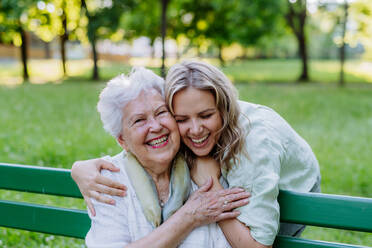 Porträt einer erwachsenen Enkelin, die ihre ältere Großmutter umarmt, während sie auf einer Parkbank sitzt. - HPIF04540