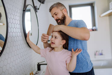 Ein Vater, der seiner kleinen Tochter im Badezimmer die Haare bürstet, Konzept der Morgenroutine. - HPIF04559