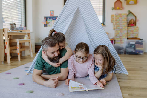 Ein fröhlicher Vater von drei kleinen Töchtern, der ihnen zu Hause ein Buch vorliest. - HPIF04581