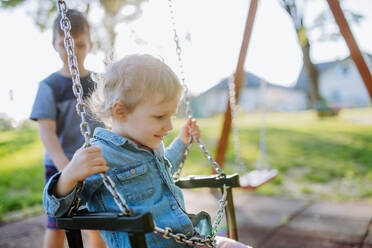 Kleine Geschwister spielen zusammen auf dem Spielplatz, schaukelnd auf einer Schaukel, genießen den sonnigen Sommertag. - HPIF04608