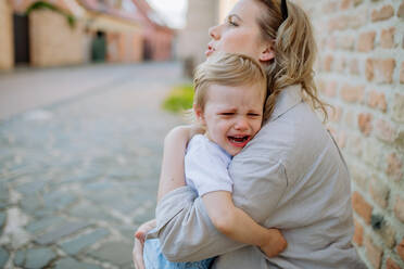 A mother consling her little daughter crying, holding her in arms in street in summer. - HPIF04615