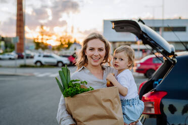 Eine junge Mutter mit ihrer kleinen Tochter hält nach dem Einkaufen abfallfreie Einkaufstüten mit Lebensmitteln in der Nähe des Autos. - HPIF04619
