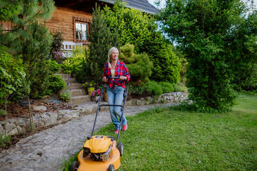 An ederly woman mowing grass with lawn mower in the garden, garden work concept. - HPIF04640