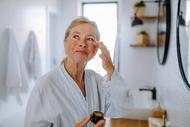 Eine schöne ältere Frau im Bademantel Anwendung natürliche Gesichtscreme im Bad, Hautpflege und Morgenroutine Konzept. - HPIF04688