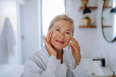 A beautiful senior woman in bathrobe, applying eye patches for puffiness while looking at camera - HPIF04696