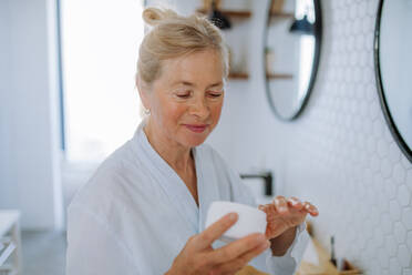 Eine schöne ältere Frau im Bademantel Anwendung natürliche Creme im Bad, Hautpflege und Morgenroutine Konzept. - HPIF04700