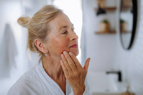 A beautiful senior woman in bathrobe looking at mirror and applying natural face cream in bathroom, skin care concept. - HPIF04701