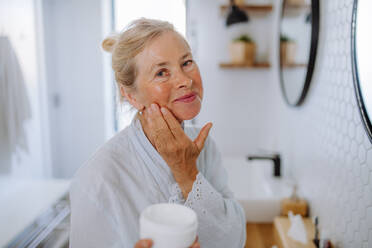 Eine schöne ältere Frau im Bademantel Anwendung natürliche Creme im Bad, Hautpflege und Morgenroutine Konzept. - HPIF04702