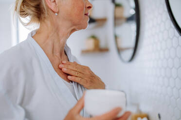 A beautiful senior woman in bathrobe applying natural cream in bathroom, skin care and morning routine concept. - HPIF04703