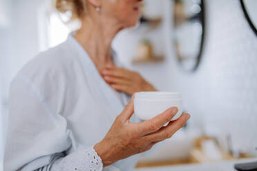 A beautiful senior woman in bathrobe applying natural cream in bathroom, skin care and morning routine concept. - HPIF04704