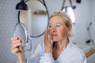 Eine schöne ältere Frau im Bademantel, die in den Spiegel schaut und natürliche Gesichtscreme im Badezimmer aufträgt, Hautpflegekonzept. - HPIF04706