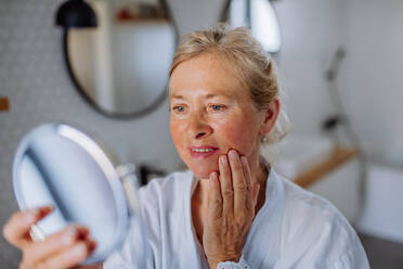 Eine schöne ältere Frau im Bademantel, die in den Spiegel schaut und natürliche Gesichtscreme im Badezimmer aufträgt, Hautpflegekonzept. - HPIF04708