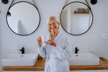 A beautiful senior woman in bathrobe drinking tea in bathroom, relax and wellness concept. - HPIF04715