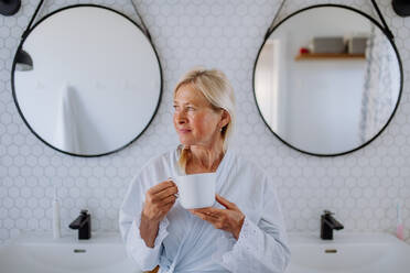 A beautiful senior woman in bathrobe drinking tea in bathroom, relax and wellness concept. - HPIF04717