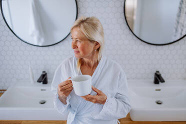 A beautiful senior woman in bathrobe drinking tea in bathroom, relax and wellness concept. - HPIF04718