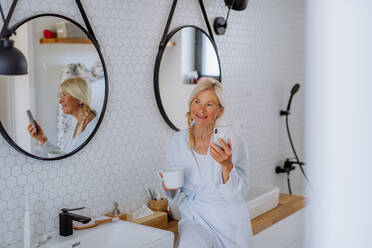 Eine schöne ältere Frau im Bademantel, die Tee trinkt und ein Smartphone im Badezimmer benutzt, Entspannungs- und Wellnesskonzept. - HPIF04719