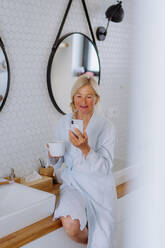 A beautiful senior woman in bathrobe drinking tea and using smartphone in bathroom, relax and wellness concept. - HPIF04720