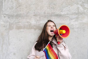 Junge Frau hält Regenbogenflagge und schreit durch ein Megaphon, das an der Wand lehnt - ASGF03202