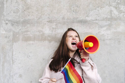Young woman holding rainbow flag shouting through megaphone leaning on wall - ASGF03202