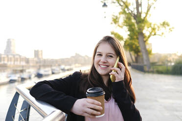 Happy young woman holding disposable cup talking on phone by railing - ASGF03208