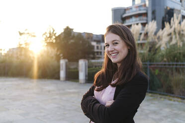 Happy young woman with arms crossed on footpath at sunset - ASGF03211