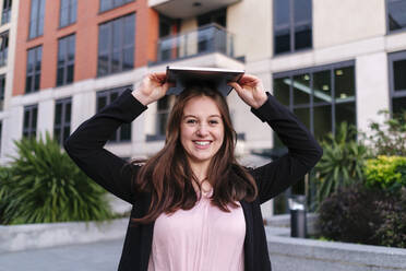 Glückliche Geschäftsfrau mit Laptop auf dem Kopf vor einem Gebäude - ASGF03216