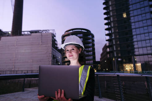 Lächelnder Ingenieur mit Laptop vor einem Gebäude in der Abenddämmerung - ASGF03218