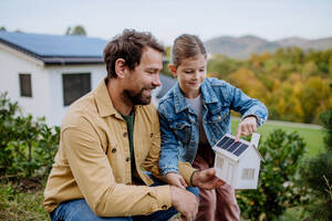 Kleines Mädchen mit ihrem Vater, der ein Papiermodell eines Hauses mit Sonnenkollektoren hält und erklärt, wie es funktioniert - alternative Energie, Ressourceneinsparung und nachhaltiger Lebensstil. - HPIF04792