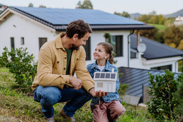 Kleines Mädchen mit ihrem Vater, der ein Papiermodell eines Hauses mit Sonnenkollektoren hält und erklärt, wie es funktioniert - alternative Energie, Ressourceneinsparung und nachhaltiger Lebensstil. - HPIF04797