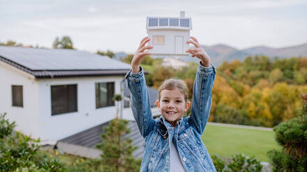 Glückliches Mädchen, das ein Papiermodell eines Hauses mit Sonnenkollektoren hält. alternative Energie, Ressourceneinsparung und nachhaltiger Lebensstil. - HPIF04831