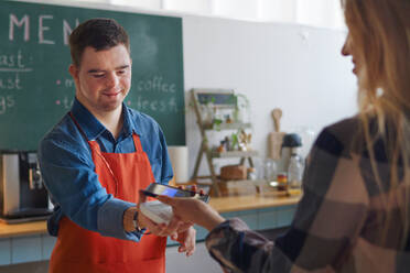 Ein fröhlicher junger Kellner mit Down-Syndrom nimmt die kontaktlose Smartphone-Zahlung eines Kunden in einem Imbissrestaurant entgegen. - HPIF04980