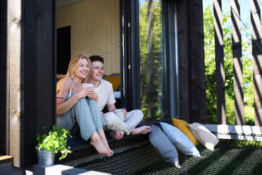 Ein junges Paar sitzt und kuschelt mit einer Tasse Kaffee auf der Terrasse ihres neuen Hauses in einem winzigen Haus im Wald, ein nachhaltiges Wohnkonzept. - HPIF05009