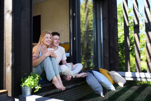 A young couple sitting and cuddling with cup of coffee in terrace in their new home in tiny house in woods, sustainable living concept. - HPIF05009