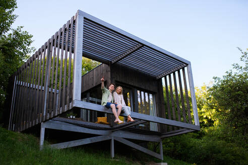 A young couple sitting and cuddling in hammock terrace in their new home in tiny house in woods, sustainable living concept. - HPIF05023