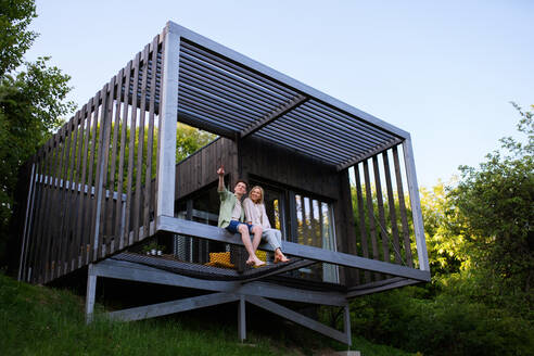 Ein junges Paar sitzt und kuschelt in der Hängematte auf der Terrasse ihres neuen Hauses in einem winzigen Haus im Wald, ein nachhaltiges Wohnkonzept. - HPIF05023