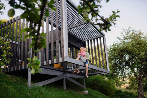Ein junges Paar sitzt und kuschelt in der Hängematte auf der Terrasse ihres neuen Hauses in einem winzigen Haus im Wald, ein nachhaltiges Wohnkonzept. - HPIF05024